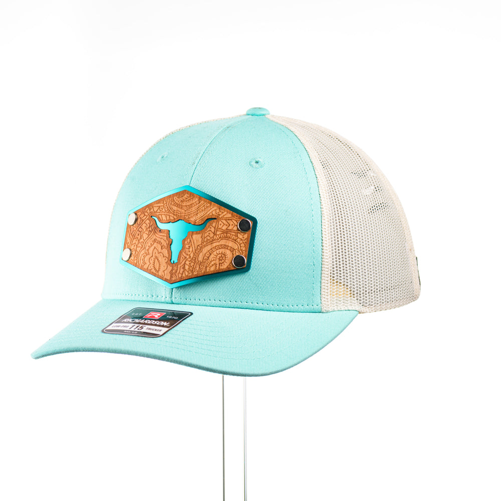 A teal and cream mesh low profile Richardson trucker hat with an etched cherry wood cut out steer skull and paisley design on a hexagon metal patch at a quarter turn.