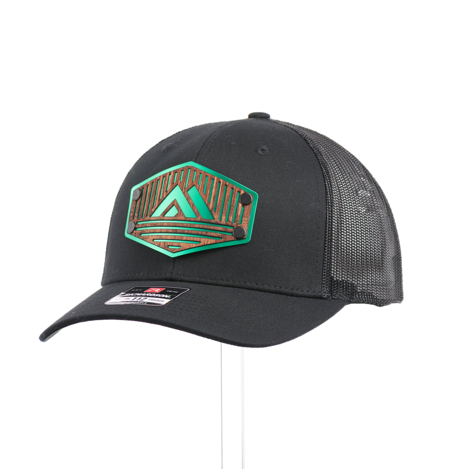 3/4 view of an Etched Mahogany Camping Triangle Patch Trucker Hat, featuring a real mahogany wood veneer patch precision-cut into a hexagon with a forest scene cut-out centered around a camping tent, laminated to a hexagonal green anodized aluminum plate, and riveted to an all-black Richardson 112 trucker hat—handmade in Montana, USA by Last Best Supply Co.