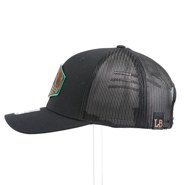 The side of an Etched Mahogany Camping Triangle Patch Trucker Hat, featuring a real mahogany wood veneer patch precision-cut into a hexagon with a forest scene cut-out centered around a camping tent, laminated to a hexagonal green anodized aluminum plate, and riveted to an all-black Richardson 112 trucker hat—handmade in Montana, USA by Last Best Supply Co.