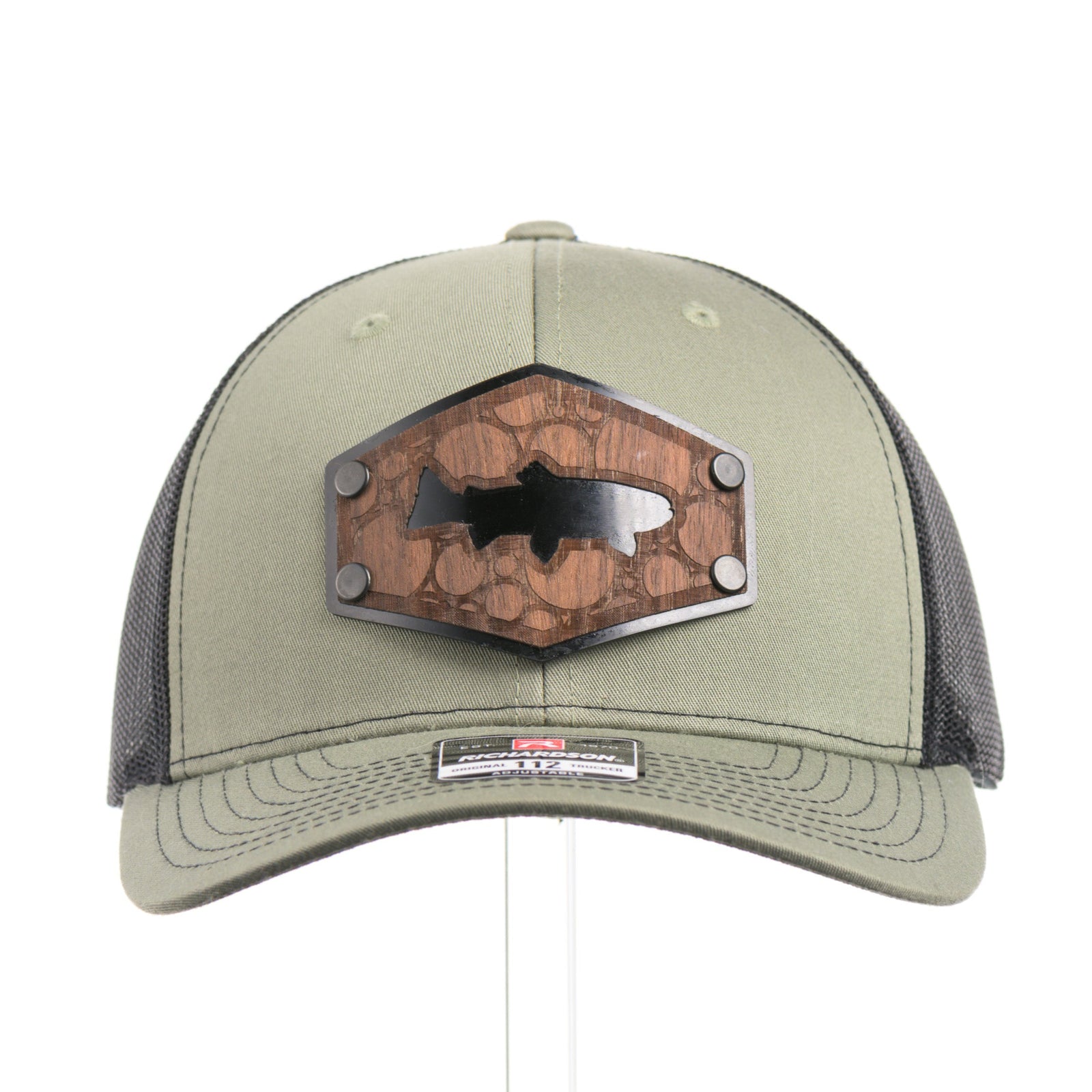 The front of a Walnut Wood Bubble Salmon Patch Trucker Hat, featuring a real walnut wood veneer patch precision-cut into a hexagon, etched with a river bubbles design with a cut-out silhouette of a salmon, laminated to a hexagonal black anodized aluminum plate, and riveted to a loden green and black mesh Richardson 112 trucker hat—handmade in Montana, USA by Last Best Supply Co.