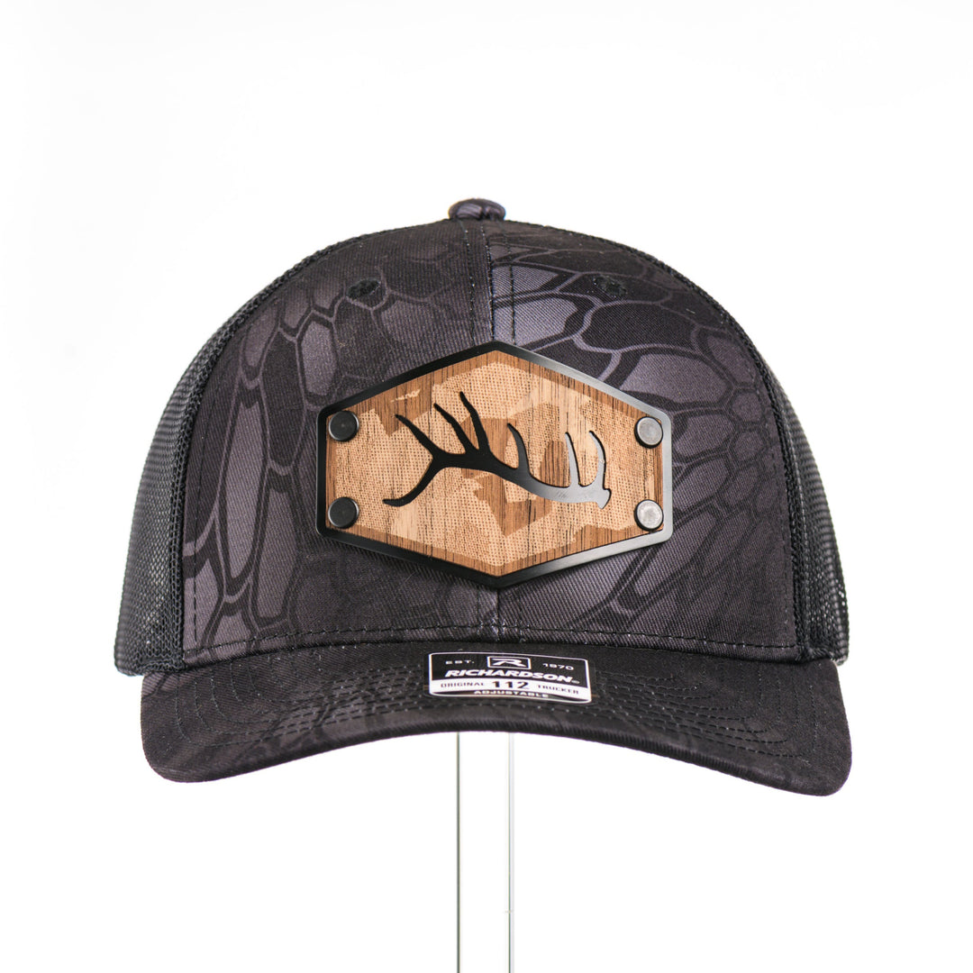 The front of a Walnut Camo Tines Kryptek Typhon Trucker Hat, featuring a hexagonal real walnut wood veneer patch with cut-out elk antler tines and laser-etched camo pattern, laminated to a hexagonal black anodized aluminum plate, and riveted to a Richardson trucker hat in Kryptek Typhon camo—handmade in Montana, USA by Last Best Supply Co.
