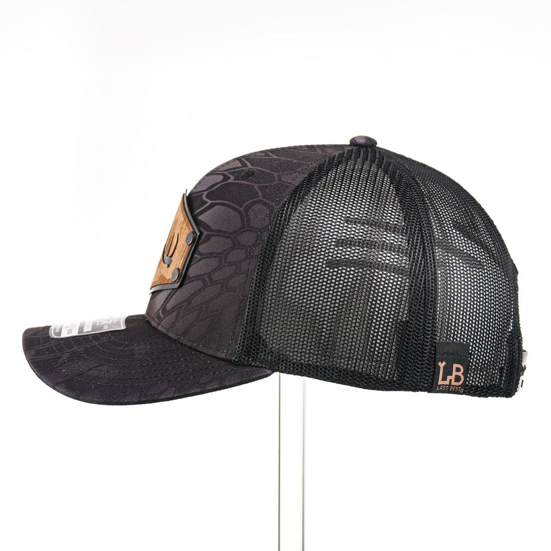 The side of a Walnut Camo Tines Kryptek Typhon Trucker Hat, featuring a hexagonal real walnut wood veneer patch with cut-out elk antler tines and laser-etched camo pattern, laminated to a hexagonal black anodized aluminum plate, and riveted to a Richardson trucker hat in Kryptek Typhon camo—handmade in Montana, USA by Last Best Supply Co.