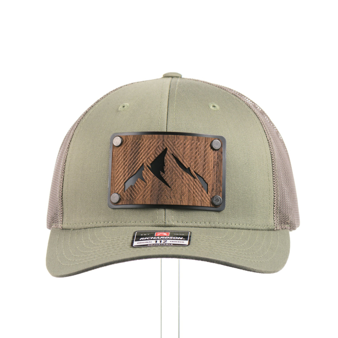 The front of an Etched Walnut Wood Mountain Patch Trucker Hat, featuring a real walnut wood patch cut into the shape of mountain peaks, mounted on a black anodized aluminum plate and secured with rivets to a loden green Richardson 112 trucker hat—handmade in Montana, USA by Last Best Supply Co.
