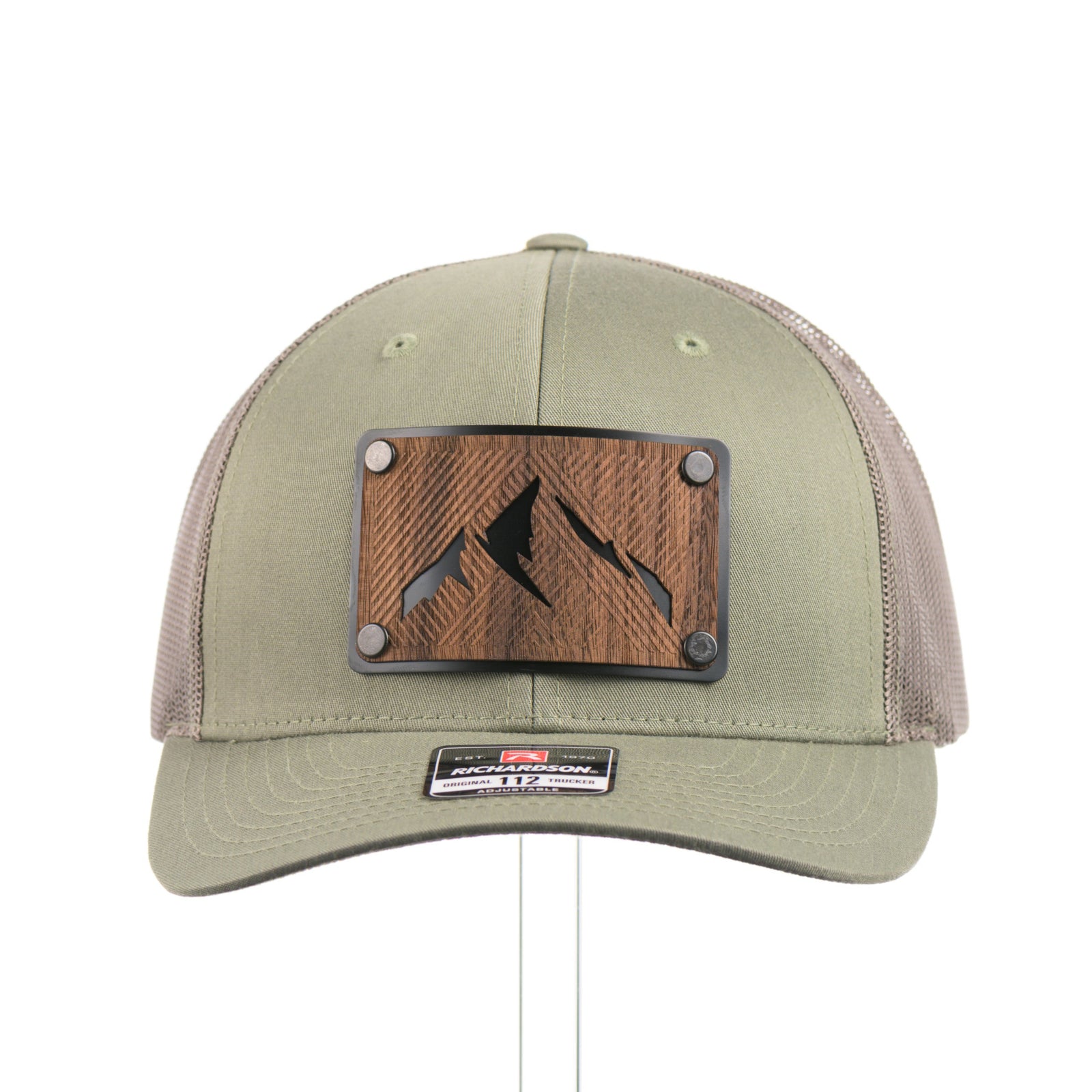 The front of an Etched Walnut Wood Mountain Patch Trucker Hat, featuring a real walnut wood patch cut into the shape of mountain peaks, mounted on a black anodized aluminum plate and secured with rivets to a loden green Richardson 112 trucker hat—handmade in Montana, USA by Last Best Supply Co.