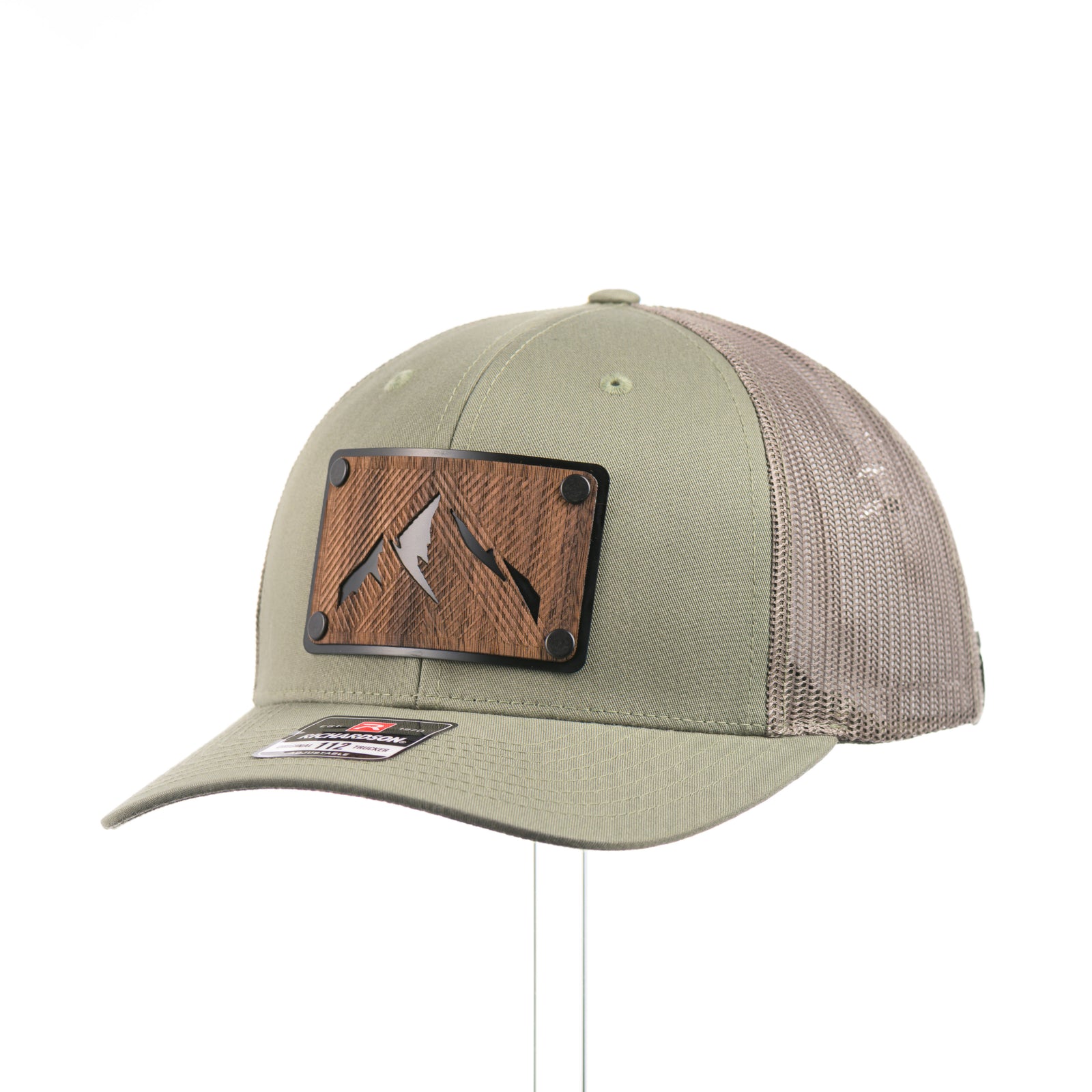 3/4 view of an Etched Walnut Wood Mountain Patch Trucker Hat, featuring a real walnut wood patch cut into the shape of mountain peaks, mounted on a black anodized aluminum plate and secured with rivets to a loden green Richardson 112 trucker hat—handmade in Montana, USA by Last Best Supply Co.