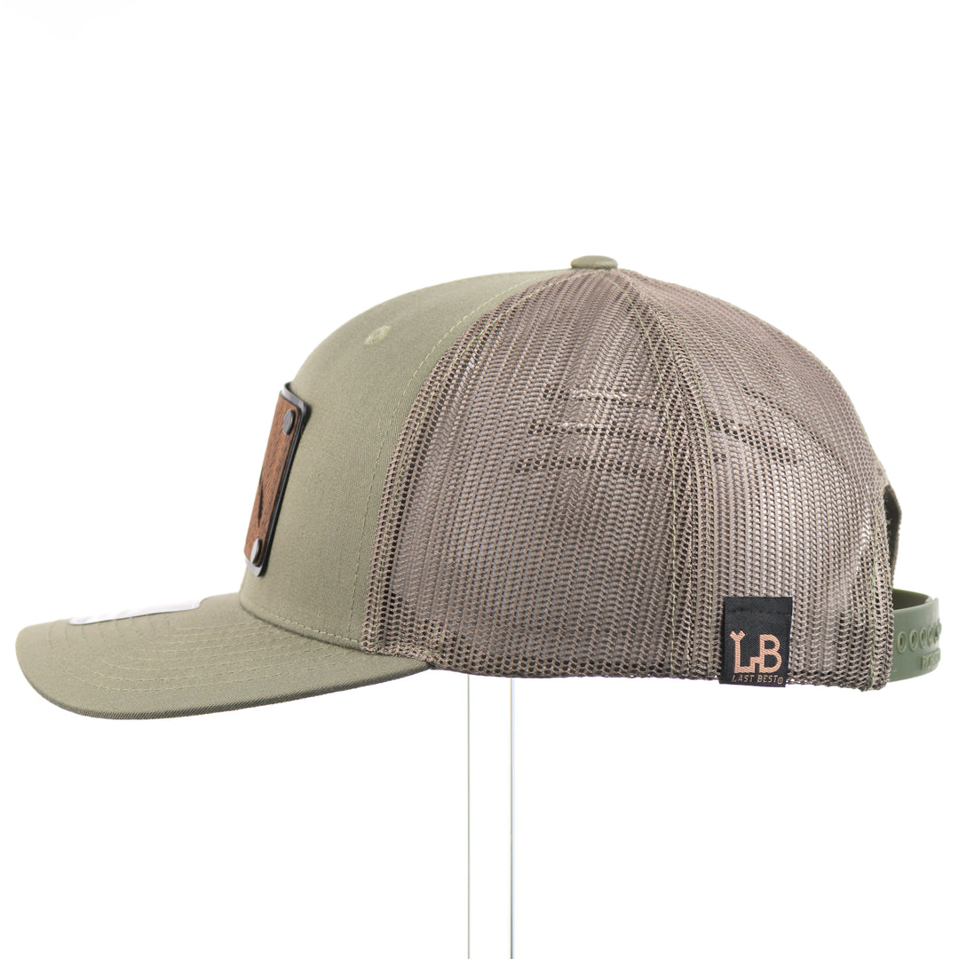 The side of an Etched Walnut Wood Mountain Patch Trucker Hat, featuring a real walnut wood patch cut into the shape of mountain peaks, mounted on a black anodized aluminum plate and secured with rivets to a loden green Richardson 112 trucker hat—handmade in Montana, USA by Last Best Supply Co.