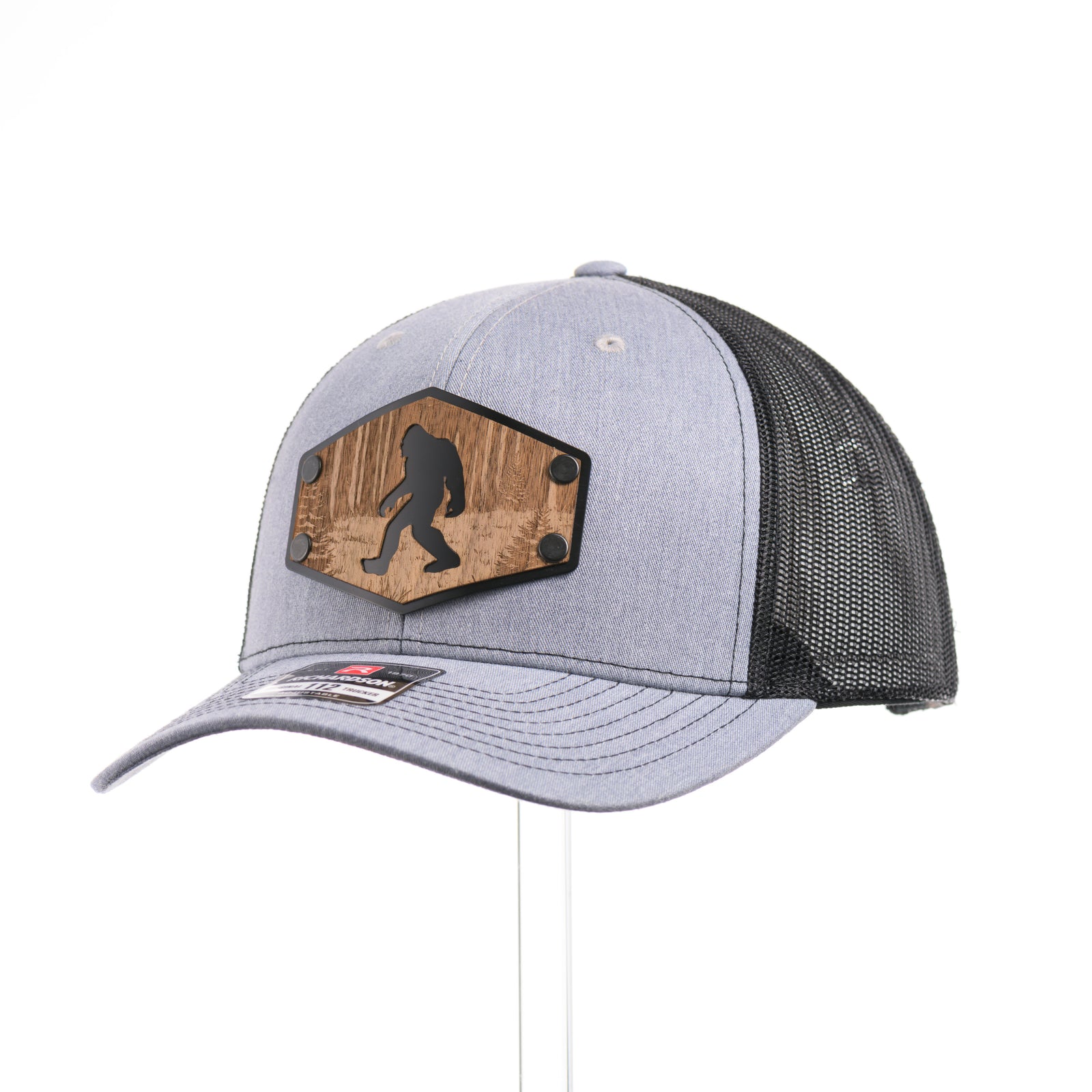 3/4 view of a Walnut Wood Roaming Bigfoot Patch Flat Bill Trucker Hat, featuring a real walnut wood veneer patch precision-cut into a hexagon, etched with a forest scene with a silhouette of a roaming Sasquatch, laminated to a hexagonal black anodized aluminum plate, and riveted to a heather gray and black mesh Richardson 112 trucker hat—handmade in Montana, USA by Last Best Supply Co.