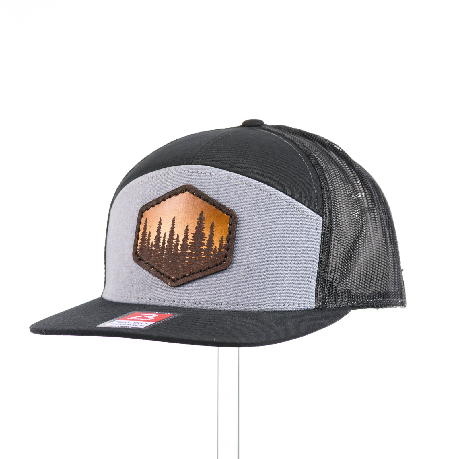 3/4 view of an Evergreen Trees Hexagon Leather Patch Flat Bill Trucker Hat, featuring a hexagonal hand-dyed leather patch etched with an evergreen forest scene, securely sewn to the structured front of a heather gray and black Richardson 168 seven-panel flat bill trucker hat—handmade in Montana, USA by Last Best Supply Co.