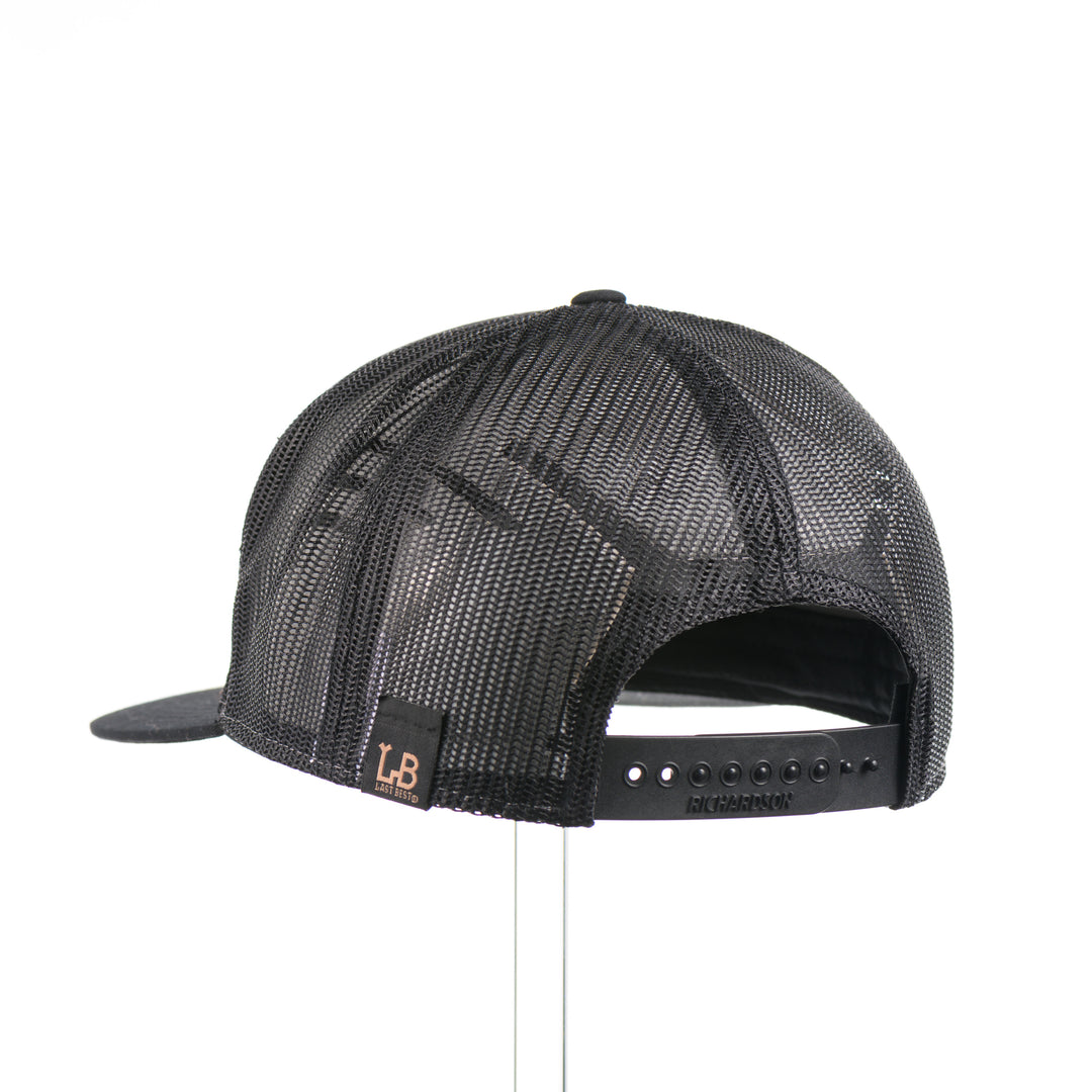 The back of an Evergreen Trees Hexagon Leather Patch Flat Bill Trucker Hat, featuring a hexagonal hand-dyed leather patch etched with an evergreen forest scene, securely sewn to the structured front of a heather gray and black Richardson 168 seven-panel flat bill trucker hat—handmade in Montana, USA by Last Best Supply Co.