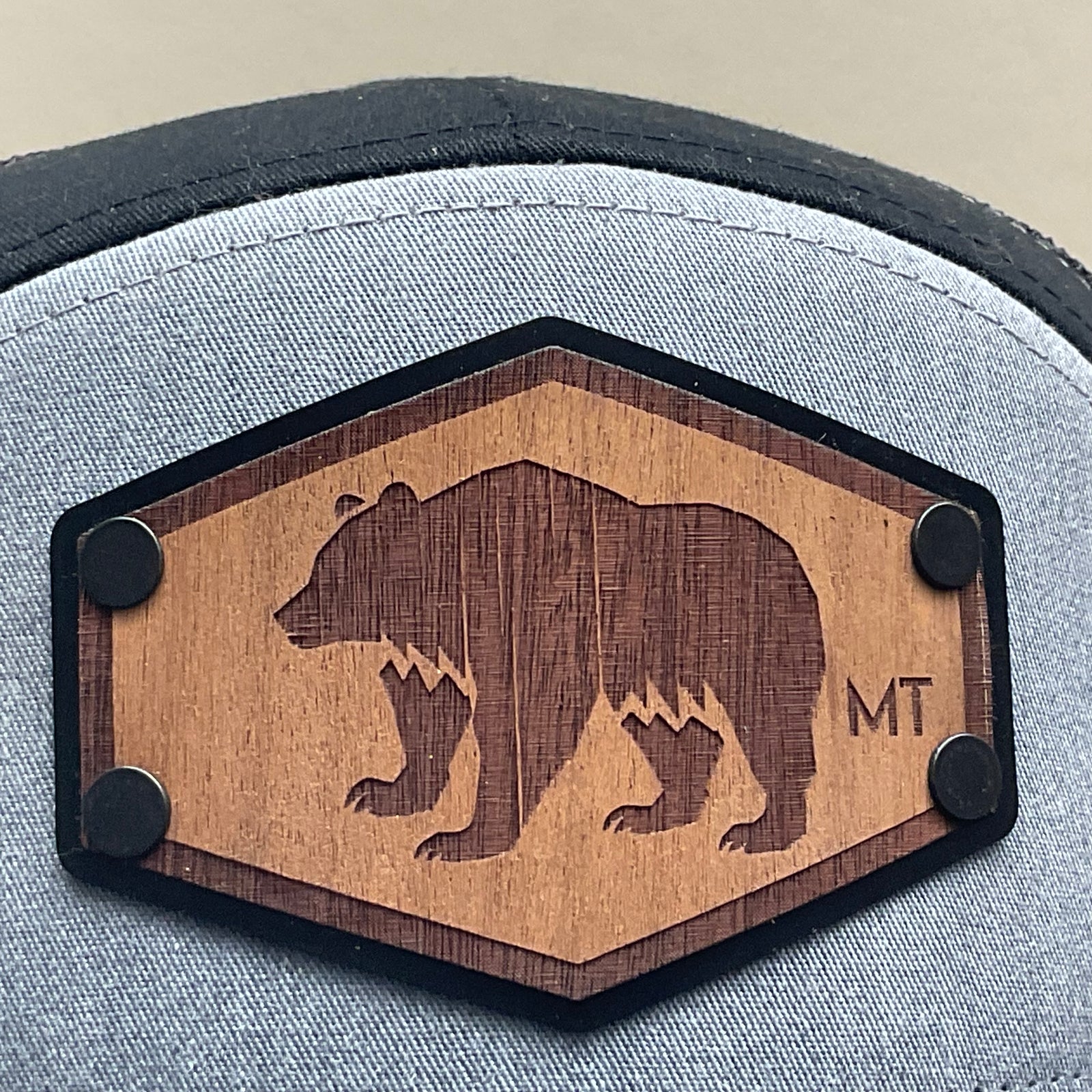 Patch detail of a Hex Bear MT Wood Patch Richardson Flat Bill Hat, featuring a hexagonal, real wood veneer patch etched with a grizzly bear and the letters “MT”, laminated to a black anodized metal plate, and then riveted to heather gray and black Richardson 168 seven-panel flat bill trucker hat—handmade in Montana, USA by Last Best Supply Co.
