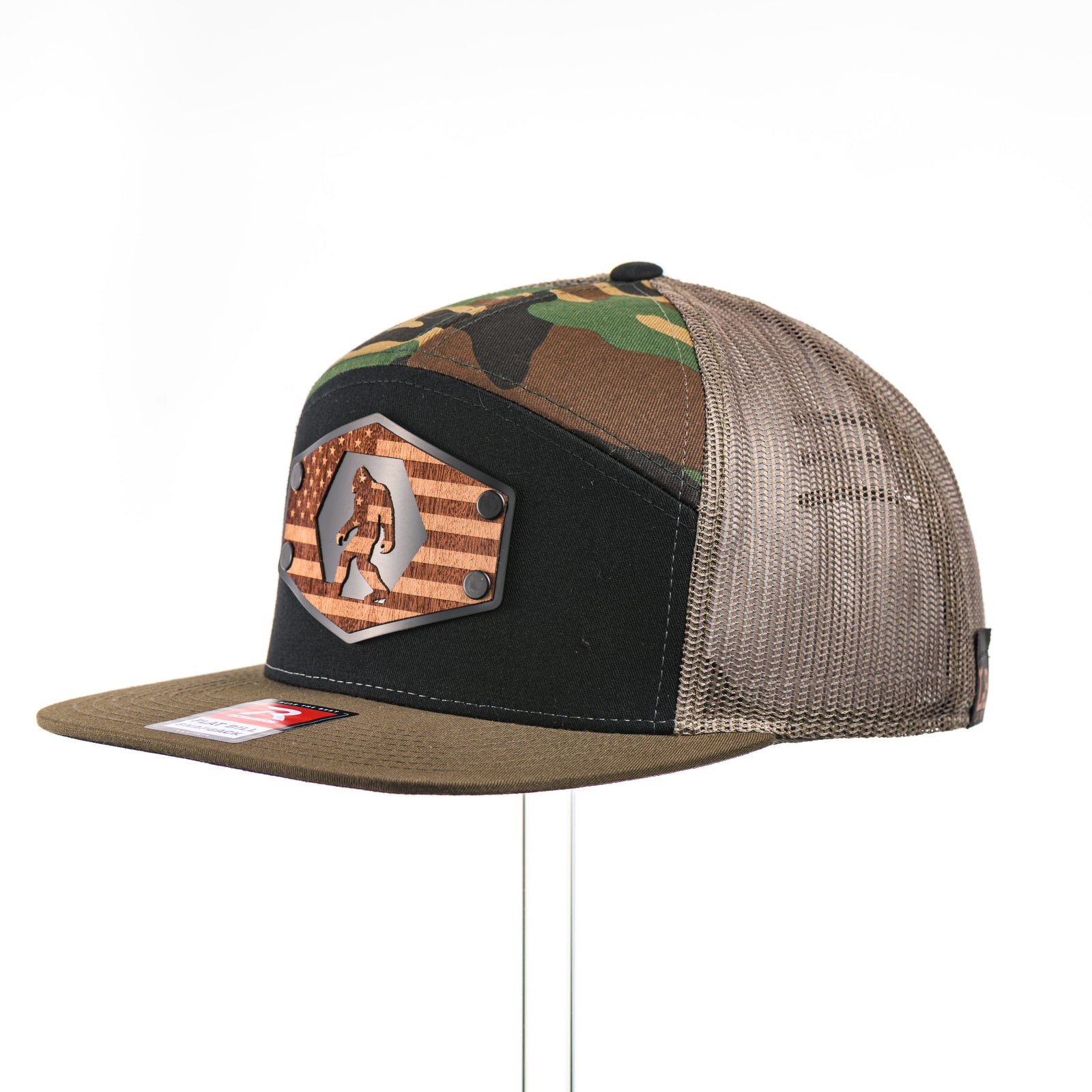 3/4 view of a Mahogany Wood Bigfoot American Flag Patch Camo Flat Bill Hat, featuring a hexagonal real mahogany wood veneer patch etched with an American flag with cut-out Bigfoot silhouette, laminated to a hexagonal black anodized aluminum plate, and riveted to a black, camo, and loden green Richardson 168 seven-panel trucker hat—handmade in Montana, USA by Last Best Supply Co.