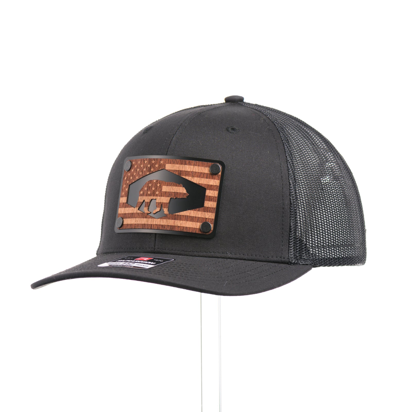 A black Richardson trucker hat with an etched mahogany wood American flag and bear cut out on a black metal patch at a quarter turn, handmade in Montana, USA by Last Best Supply Co.