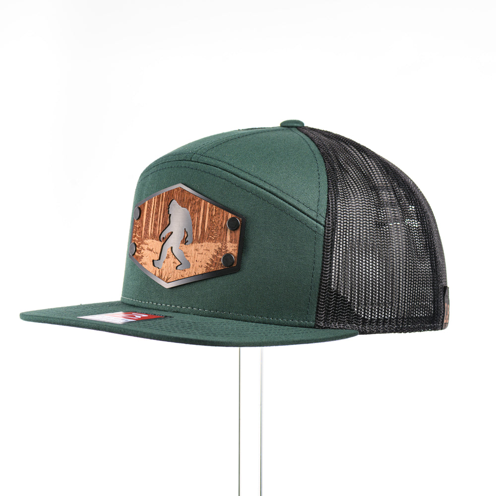 A dark green flat bill trucker hat with an etched mahogany wood roaming Sasquatch patch on a hexagon metal plate at a quarter turn, made in Montana USA by Last Best Supply Co.