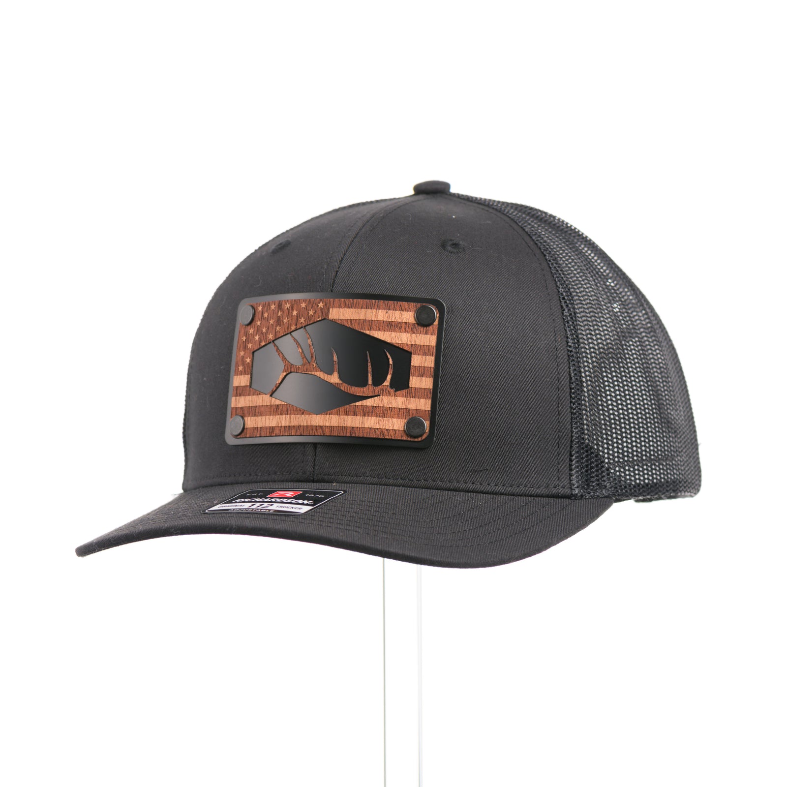 3/4 view of a Mahogany Wood Tines American Flag Patch Trucker Hat, featuring a real walnut wood veneer patch etched with an American flag and cut-out elk antler tines, laminated to a black anodized aluminum plate and riveted to an all-black Richardson 112 trucker hat along—handmade in Montana, USA by Last Best Supply Co.