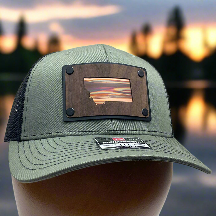 The front of a Montana Walnut Wood with Sundae Copper Patch on Richardson Trucker Hat, featuring a rectangular, real walnut wood veneer patch with a precision-cut state of Montana silhouette cutout, laminated to a black anodized metal plate, and then riveted to an olive green Richardson 112 trucker hat with black mesh—handmade in Montana, USA by Last Best Supply Co. (with a sunset over the lake background)