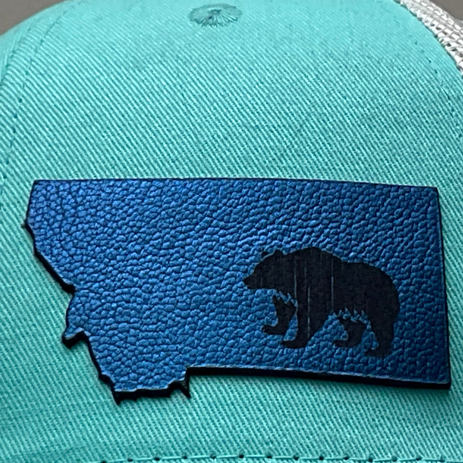 Patch detail of a Premium Leather Montana Bear on Low Profile Teal Trucker Hat, featuring a blue hand-dyed leather patch cut in the shape of the state of Montana with a grizzly bear cut-out, heat pressed to the structured front of a teal and white  trucker hat—handmade in Montana, USA by Last Best Supply Co.