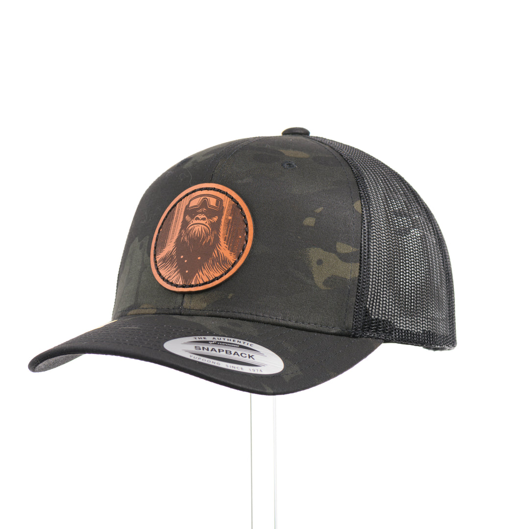3/4 view of a Snowy Bigfoot Leather Patch Black Camo Trucker Hat, featuring a circular real hand-dyed leather patch etched with a detailed Bigfoot in ski googles design, securely heat bonded and sewn onto the structured front of a dark camo Yupoong Retro Trucker Hat—handmade in Montana, USA by Last Best Supply Co.