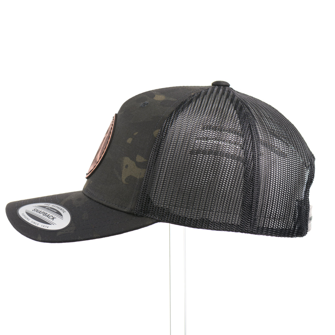 The side of a Snowy Bigfoot Leather Patch Black Camo Trucker Hat, featuring a circular real hand-dyed leather patch etched with a detailed Bigfoot in ski googles design, securely heat bonded and sewn onto the structured front of a dark camo Yupoong Retro Trucker Hat—handmade in Montana, USA by Last Best Supply Co.
