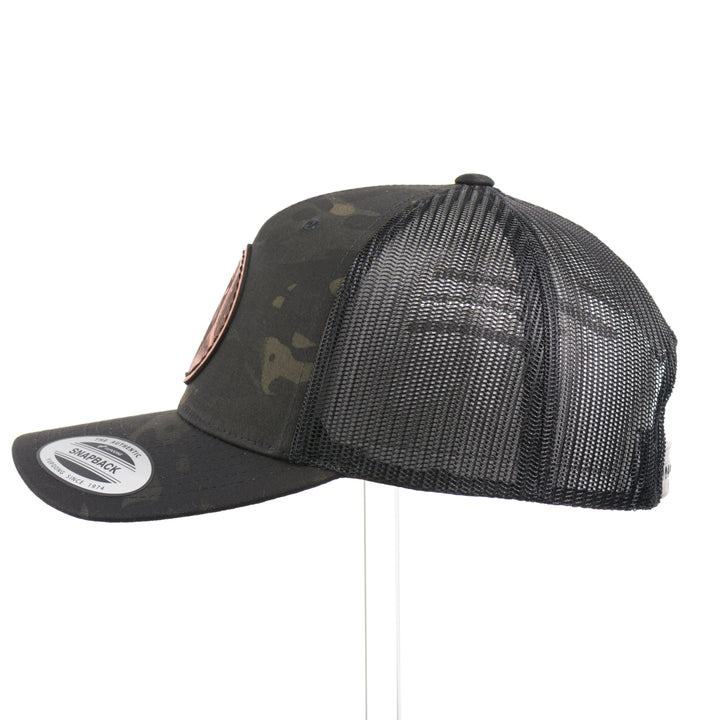 The side of a Snowy Bigfoot Leather Patch Black Camo Trucker Hat, featuring a circular real hand-dyed leather patch etched with a detailed Bigfoot in ski googles design, securely heat bonded and sewn onto the structured front of a dark camo Yupoong Retro Trucker Hat—handmade in Montana, USA by Last Best Supply Co.