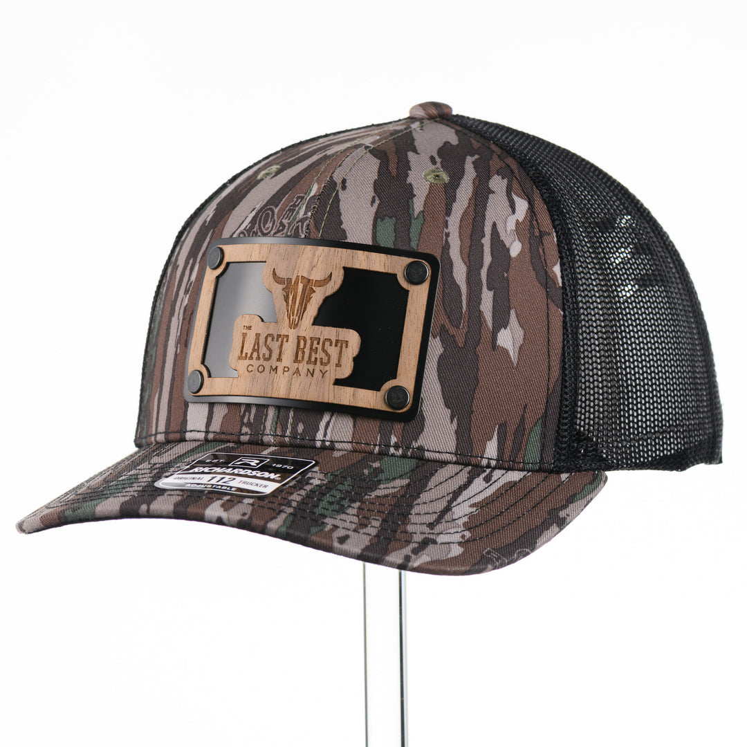 The Last Best Company Original Wood Patch Hat on Realtree Camo Richardson Hat, featuring a real wood veneer patch of The Last Best Company logo riveted to a camo trucker hat along with a metal plate—handmade in Montana, USA by Last Best Supply Co.