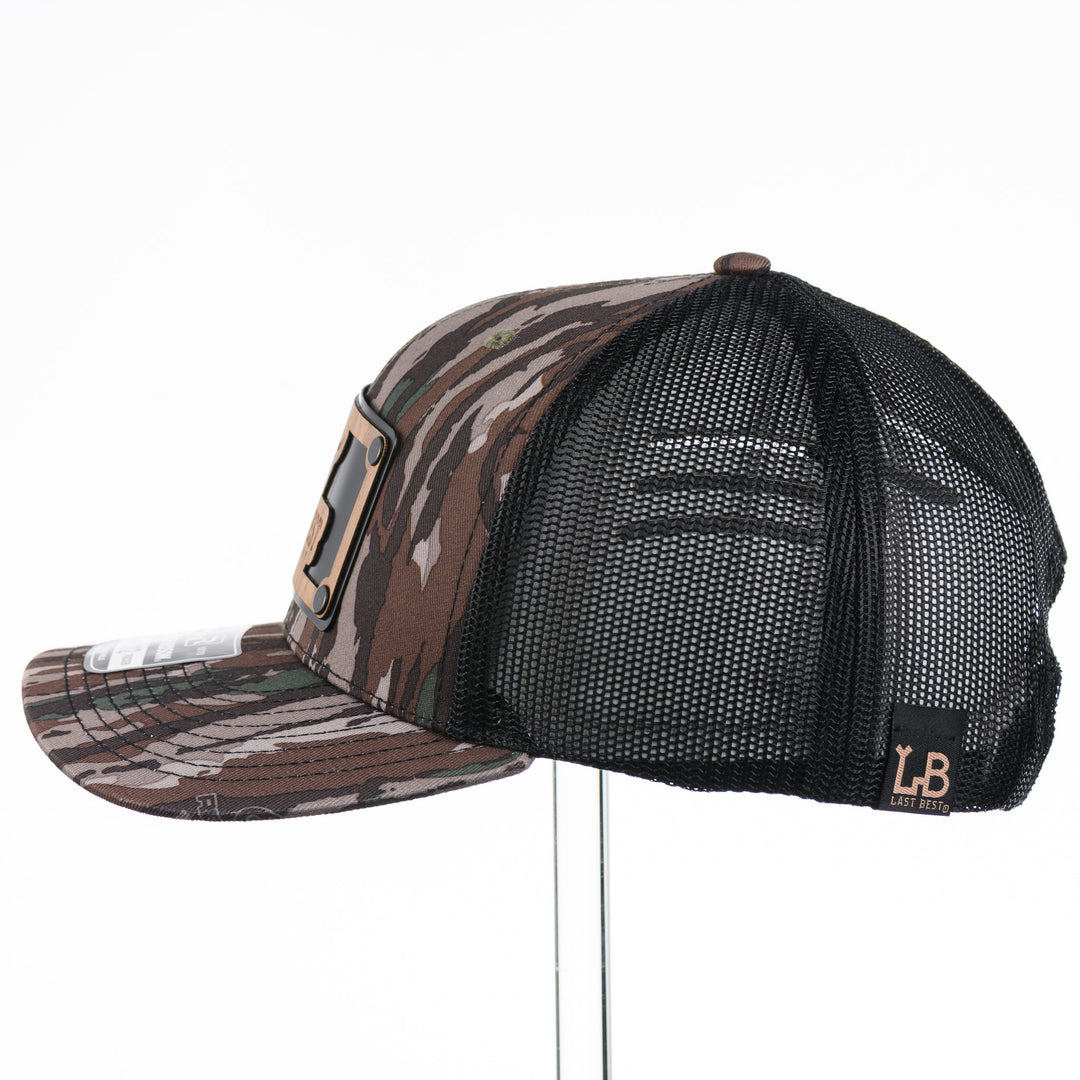 The Last Best Company Original Wood Patch Hat on Realtree Camo Richardson Hat, featuring a real wood veneer patch of The Last Best Company logo riveted to a camo trucker hat along with a metal plate—handmade in Montana, USA by Last Best Supply Co.