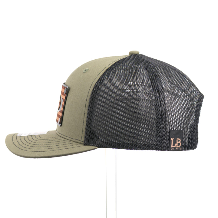The side of a loden green and black mesh Richardson trucker hat with an etched  walnut wood American flag and bison cut out on a black metal patch—handmade in Montana, USA by Last Best Supply Co.