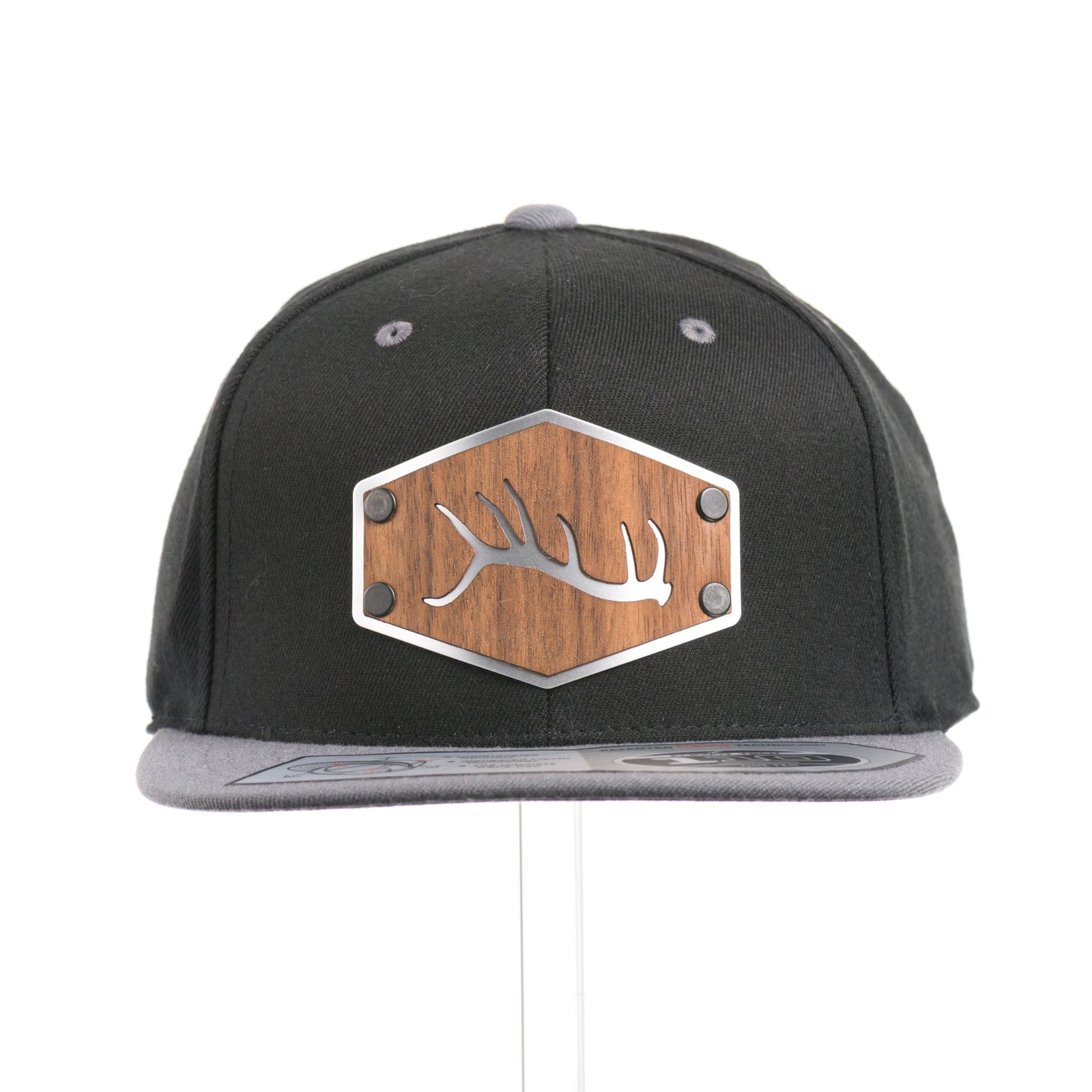 The front of a Walnut Wood Tines Flexfit Black & Grey Flat Bill Hat, featuring a real walnut wood veneer patch precision-cut into a hexagon with bold cut-out elk antler tines, laminated to a hexagon silver anodized aluminum plate, and riveted to a heather gray and black Flexfit 110F trucker hat—handmade in Montana, USA by Last Best Supply Co.