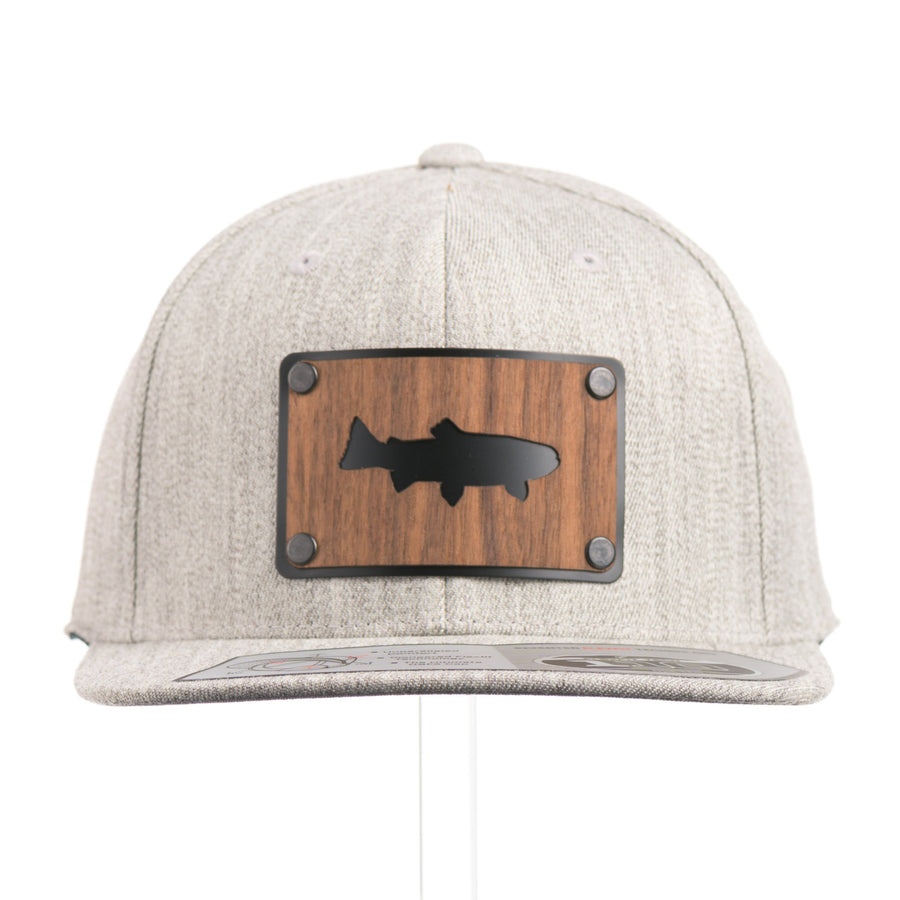 The front of a Walnut Trout Patch Plate Heather Grey Flat Bill Hat, featuring a rectangular real walnut wood veneer patch with cut-out trout silhouette, laminated to a black anodized metal plate, and riveted to a light heather grey 6-panel Flexfit Flat Bill Hat—handmade in Montana, USA by Last Best Supply Co.