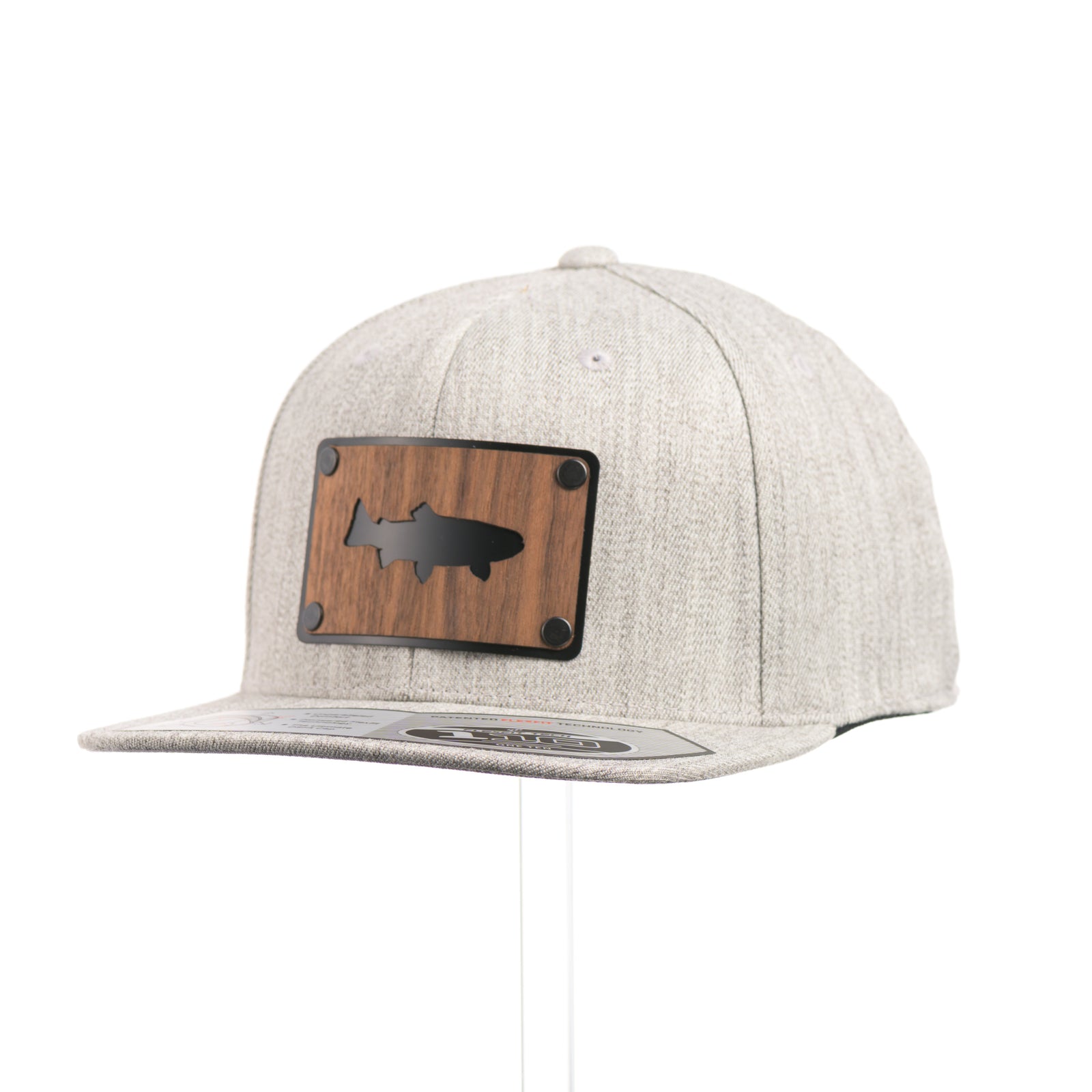 3/4 view of a Walnut Trout Patch Plate Heather Grey Flat Bill Hat, featuring a rectangular real walnut wood veneer patch with cut-out trout silhouette, laminated to a black anodized metal plate, and riveted to a light heather grey 6-panel Flexfit Flat Bill Hat—handmade in Montana, USA by Last Best Supply Co.