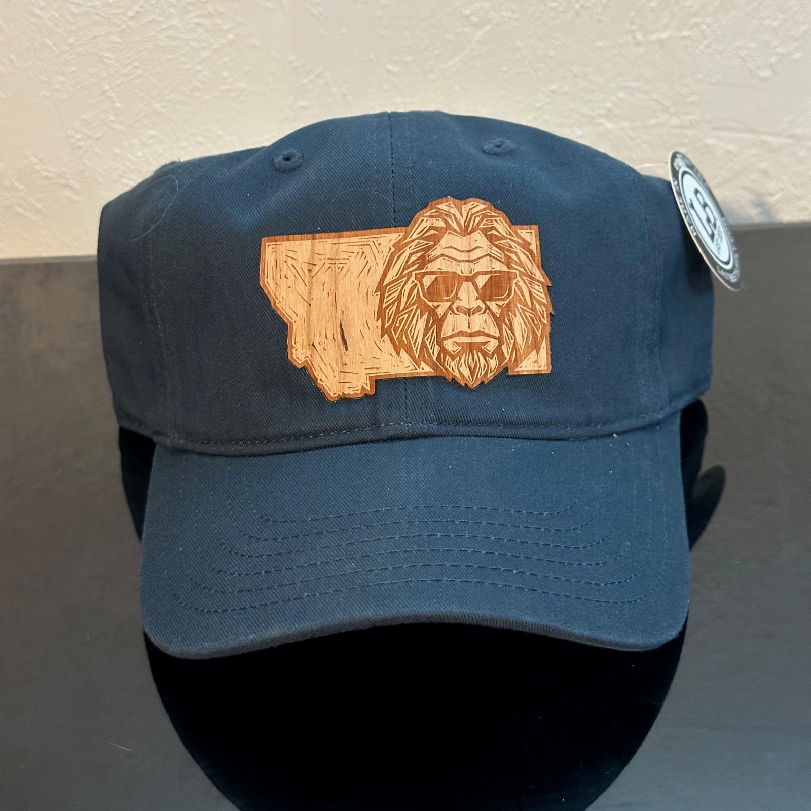 Front of an EConscious Navy 100% Cotton Unstructured Baseball Hat
decorated with a Montana Bigfoot Cherry Wood Patch - Bigfoot’s head wearing sunglasses, superimposed on the state of Montana - made in Montana by Last Best Supply Co.
