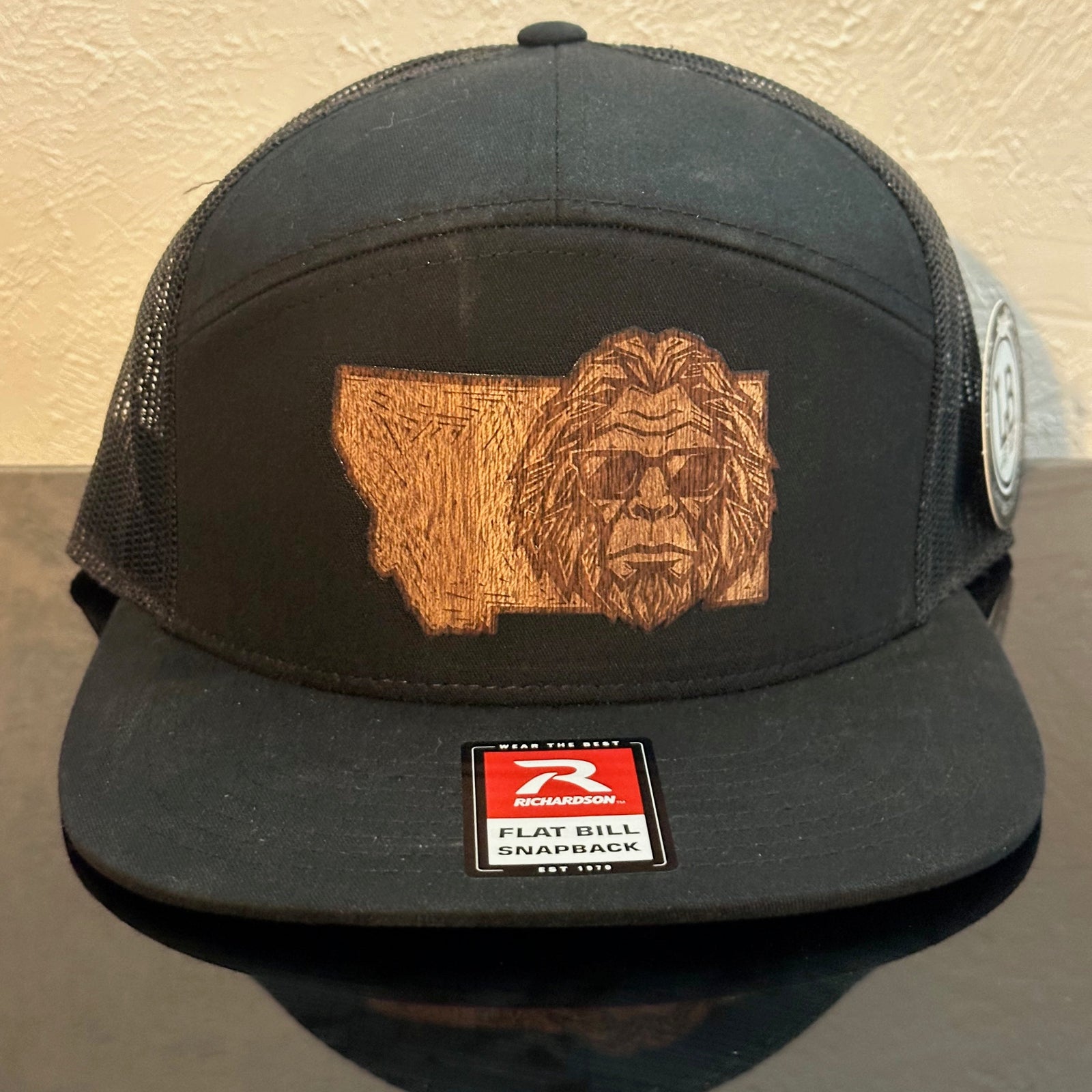 Front of a black Richardson 168 Black 7-Panel trucker hat decorated with a Montana Bigfoot Mahogany Wood Patch - Bigfoot’s head with sunglasses superimposed on the state of Montana - made in Montana by Last Best Supply Co.
