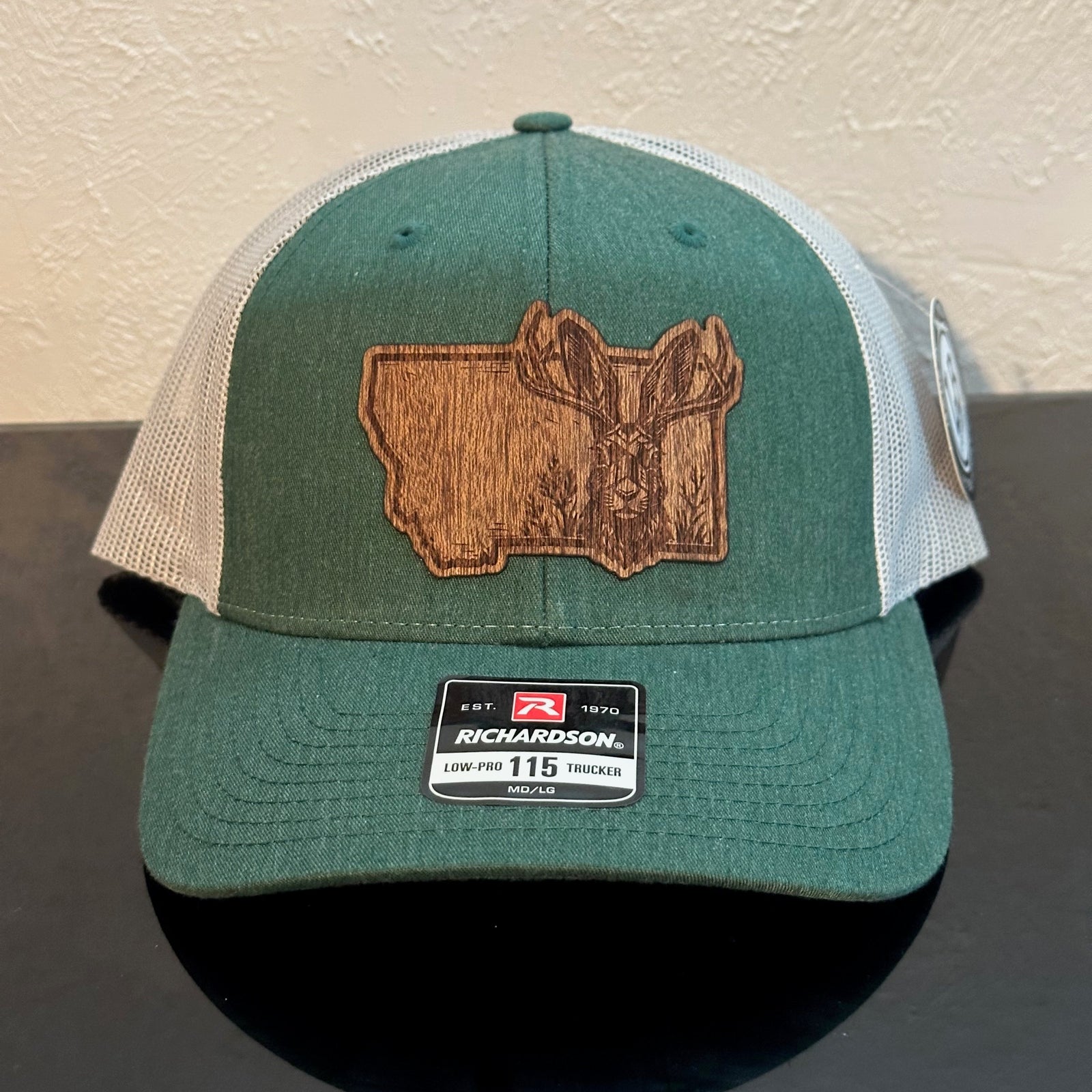 Front of a Richardson 115 Heather Green trucker hat decorated with a Montana Jackalope Mahogany Wood Patch - the head of a jackalope & wildflowers superimposed on the state of Montana - made in Montana by Last Best Supply Co.