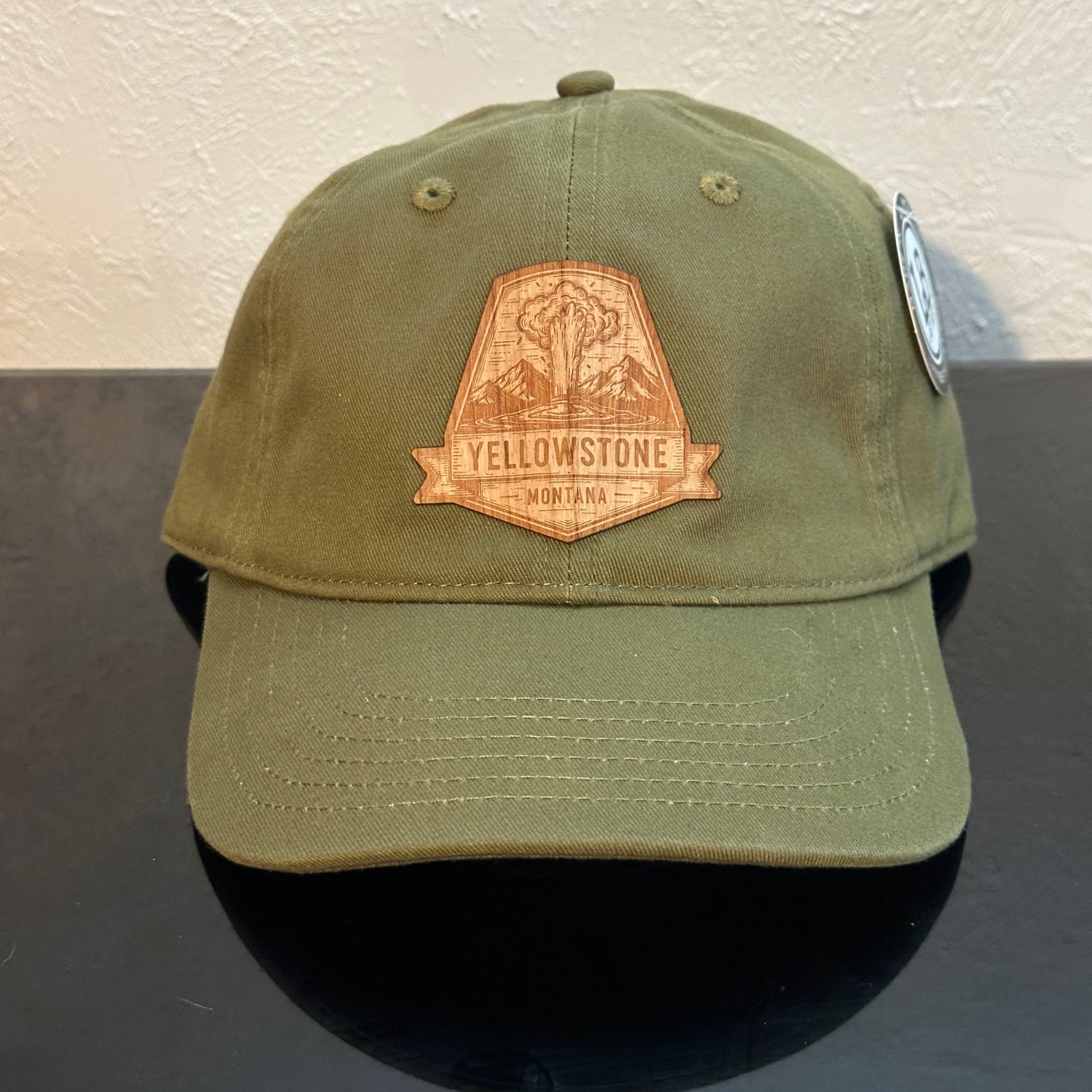 Front of an EConscious Olive Green 100% Cotton Unstructured Baseball Hat decorated with a Yellowstone Montana Wood Patch - a sketch of an erupting geyser, above the text ‘Yellowstone Montana’ - made in Montana by Last Best Supply Co.