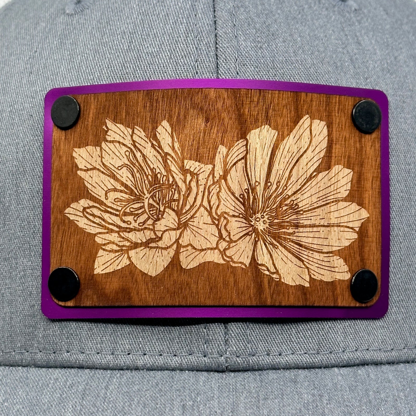 Patch detail of a Bitterroot Cherry Wood Patch Grey & White Trucker Hat, featuring a rectangular real cherry wood veneer patch with an intricate etched bitterroot flower design, laminated to a purple anodized metal plate, and riveted to a grey and white mesh trucker hat—handmade in Montana, USA by Last Best Supply Co.