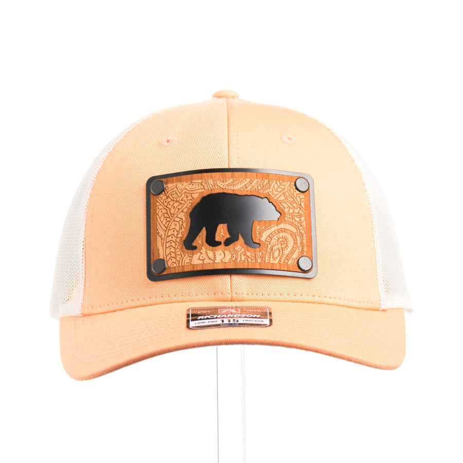 The front of a peach and birch mesh Richardson low profile trucker hat with an etched cherry wood bear cut out with an etched paisley design on a black metal plate.