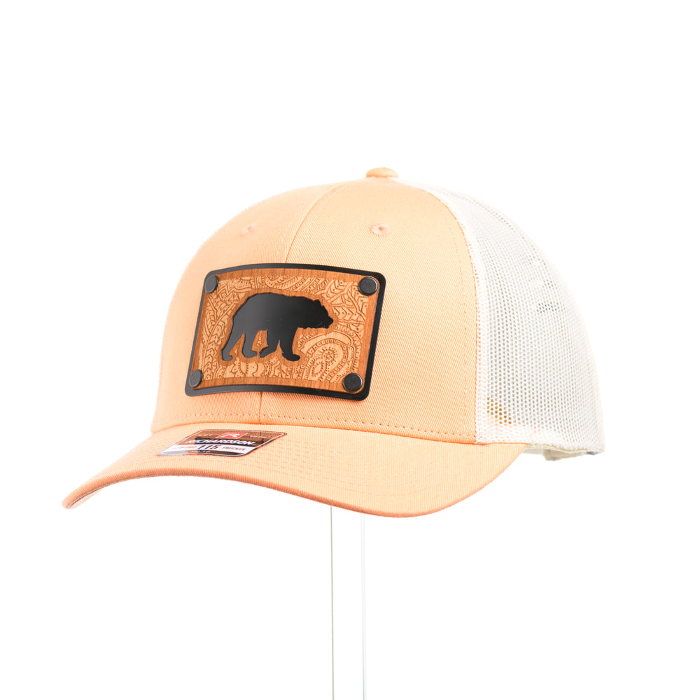 A peach and birch mesh Richardson low profile trucker hat with an etched cherry wood paisley design and bear cut out on a black metal plate at a quarter turn.