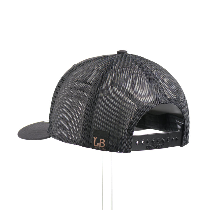 The back of a black Richardson trucker hat with an etched mahogany wood American flag and bear cut out on a black metal patch.