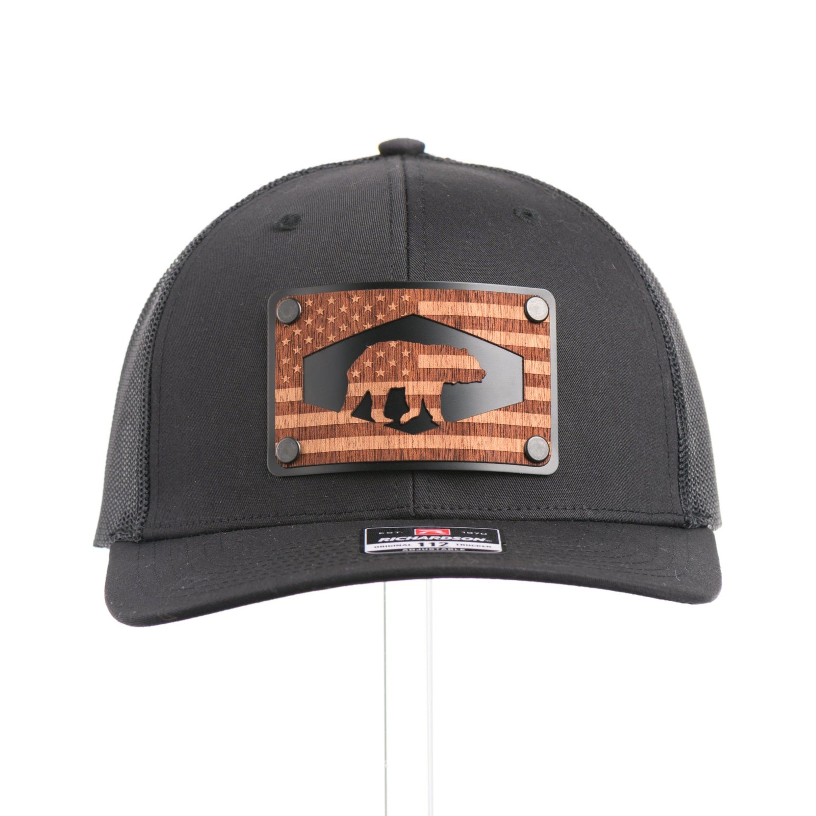 The front of a black Richardson trucker hat with an etched mahogany wood American flag and bear cut out on a black metal patch.