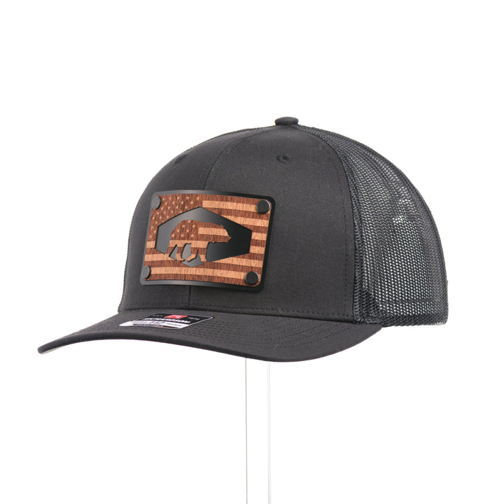 A black Richardson trucker hat with an etched mahogany wood American flag and bear cut out on a black metal patch at a quarter turn.