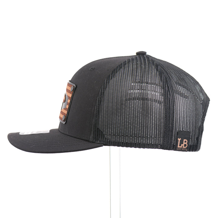 The side of a black Richardson trucker hat with an etched mahogany wood American flag and bear cut out on a black metal patch.