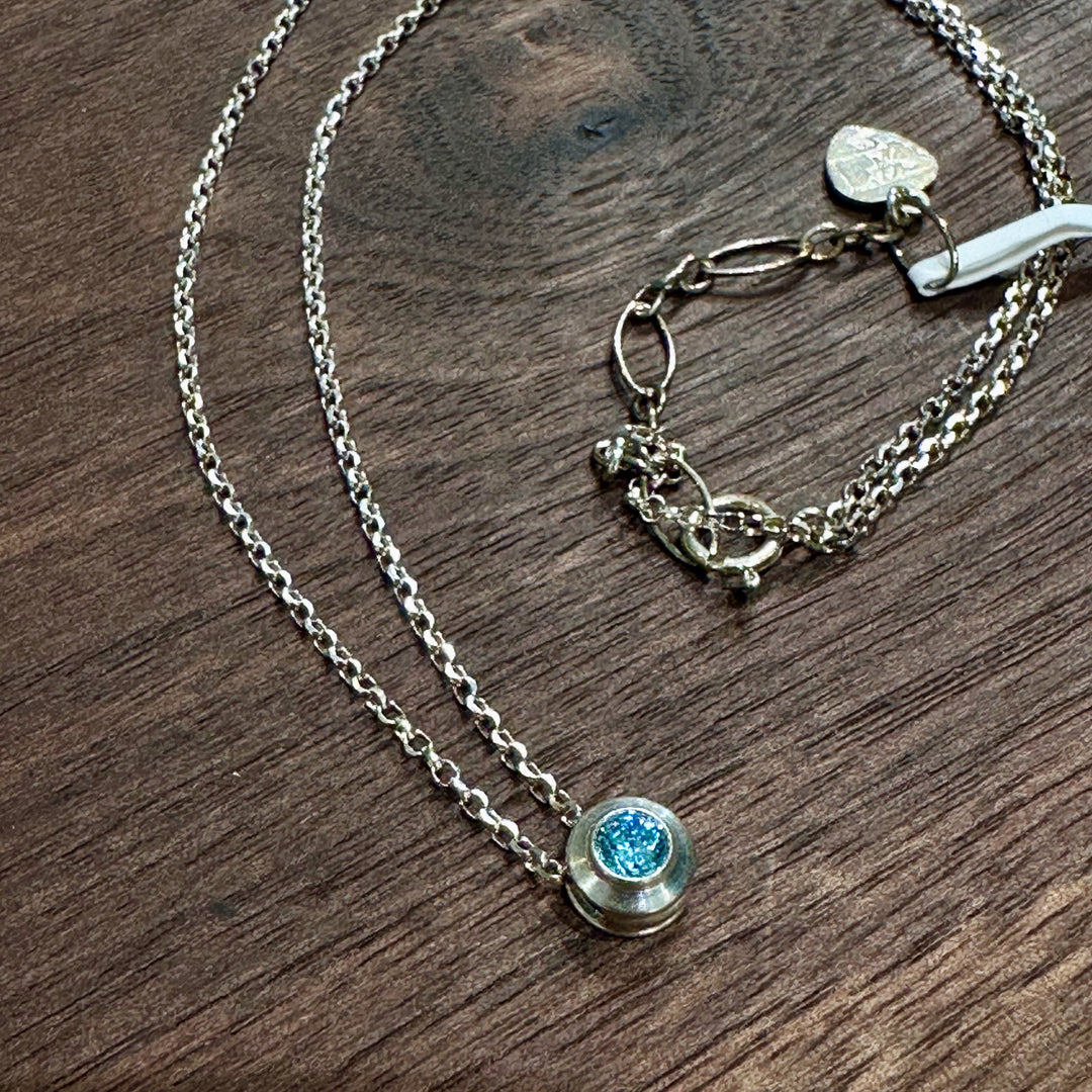 A cubic zirconia gemstone colored brilliant blue Aquamarine for a MarchBirthstone Necklace, set in sterling silver on a dark wood background