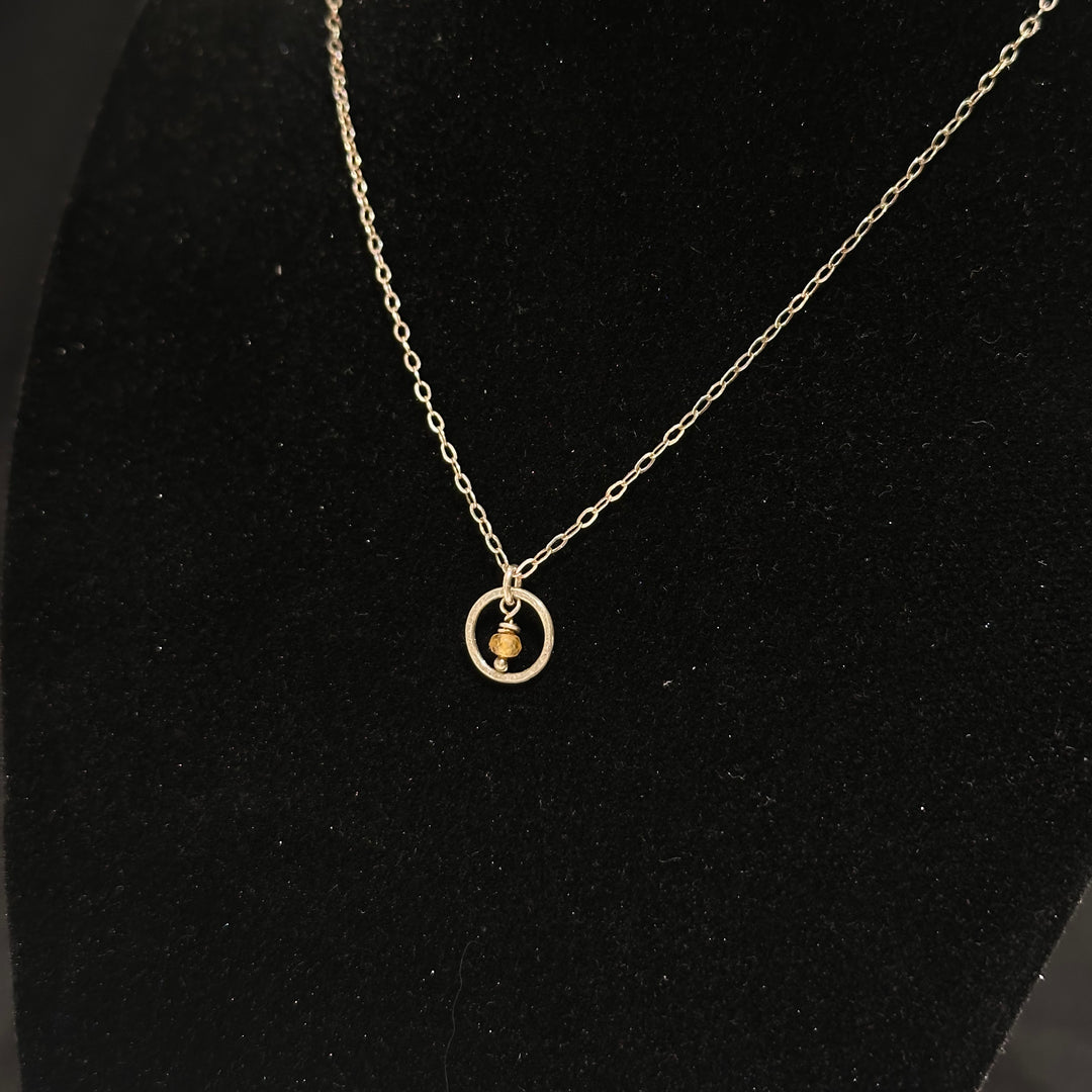 A sterling silver necklace with a small circle with a Montana sapphire bead in the center on a silver chain. Displayed on a black background