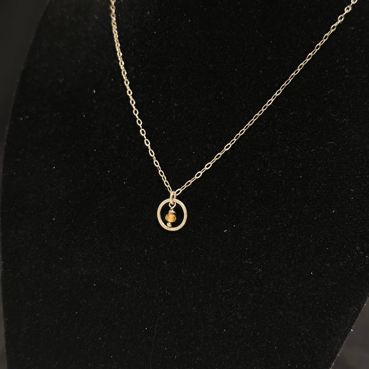 A sterling silver necklace with a small circle with a Montana sapphire bead in the center on a silver chain. Displayed on a black background