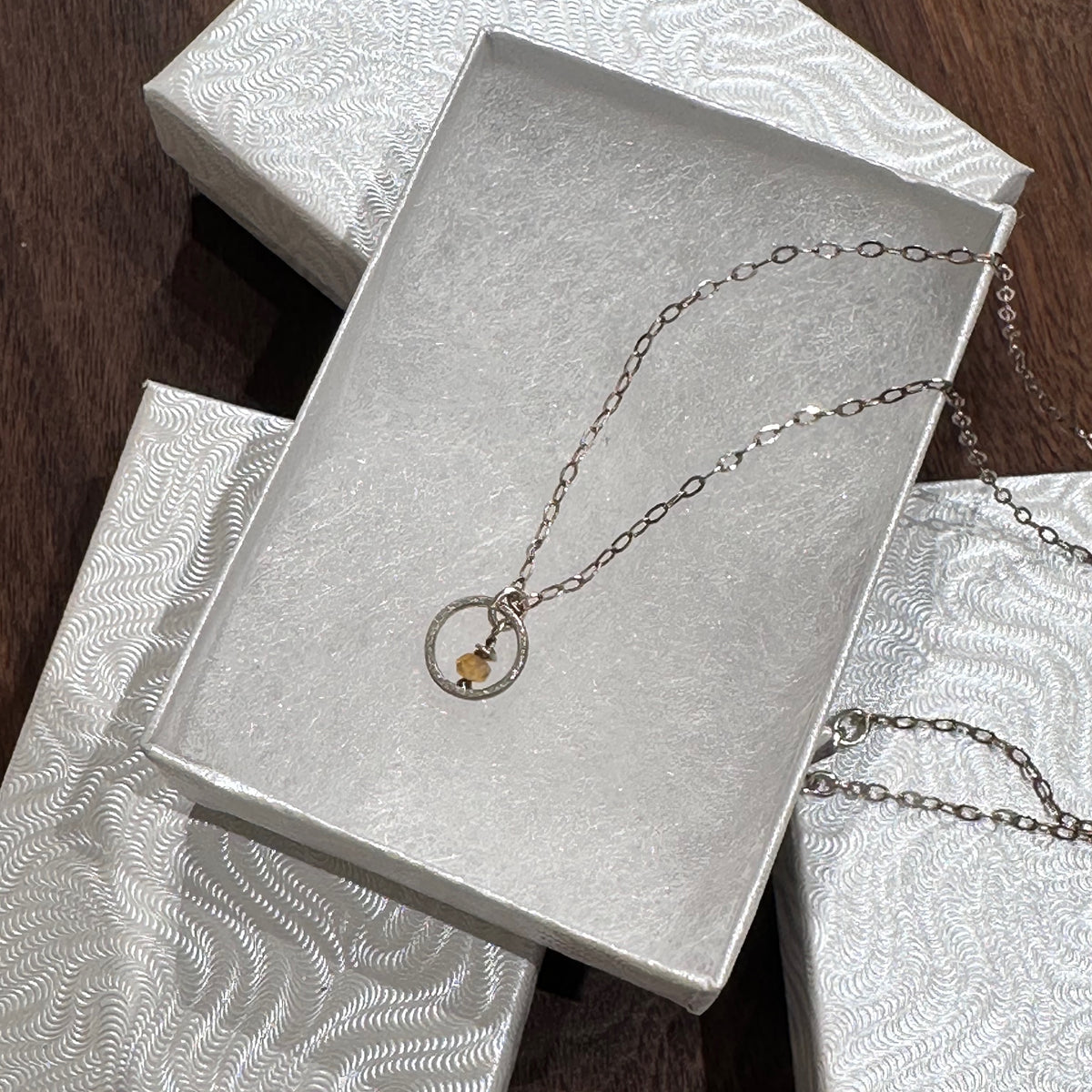 A sterling silver necklace with a small circle with a Montana sapphire bead in the center on a silver chain. Displayed in a white jewelry box on a dark wood background.