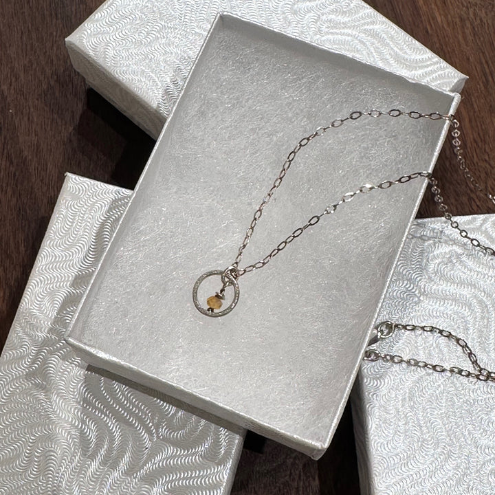 A sterling silver necklace with a small circle with a Montana sapphire bead in the center on a silver chain. Displayed in a white jewelry box on a dark wood background.