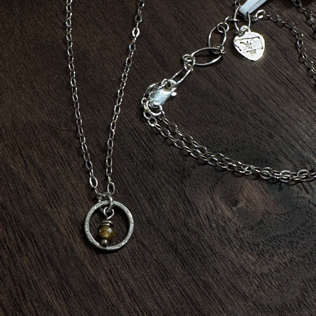 A sterling silver necklace with a small circle with a Montana sapphire bead in the center on a silver chain. Displayed in on a dark wood background