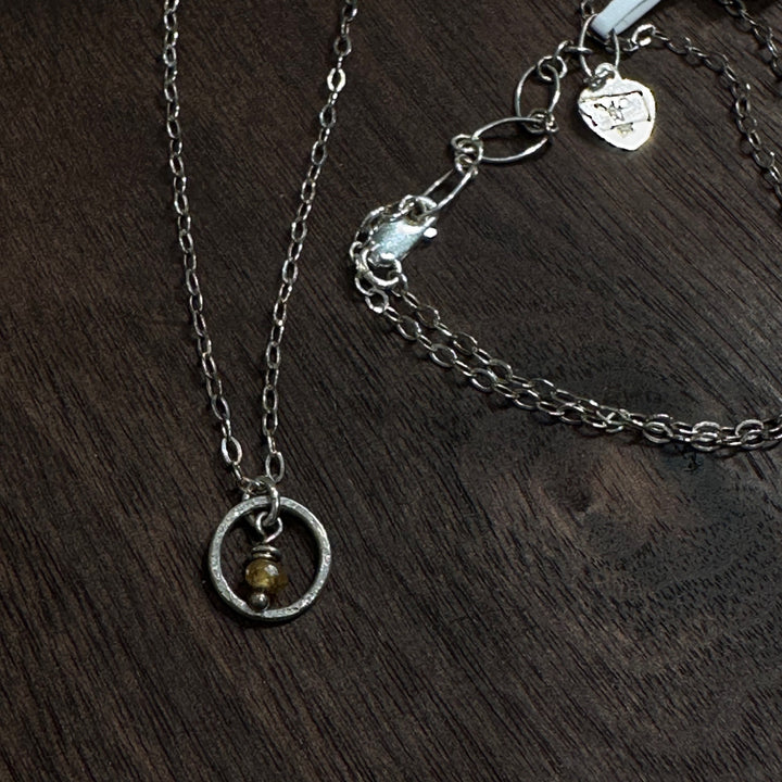 A sterling silver necklace with a small circle with a Montana sapphire bead in the center on a silver chain. Displayed in on a dark wood background