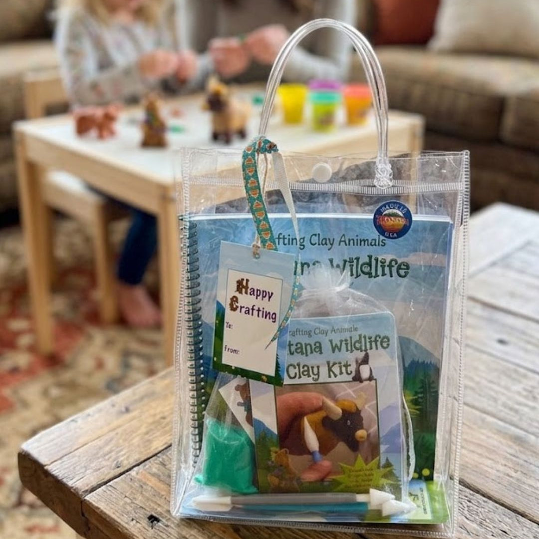 A clear plastic tote bag filled with a clay kit and booklet, placed on a wooden table in a living room. A child and an adult are blurred in the background at a craft table.