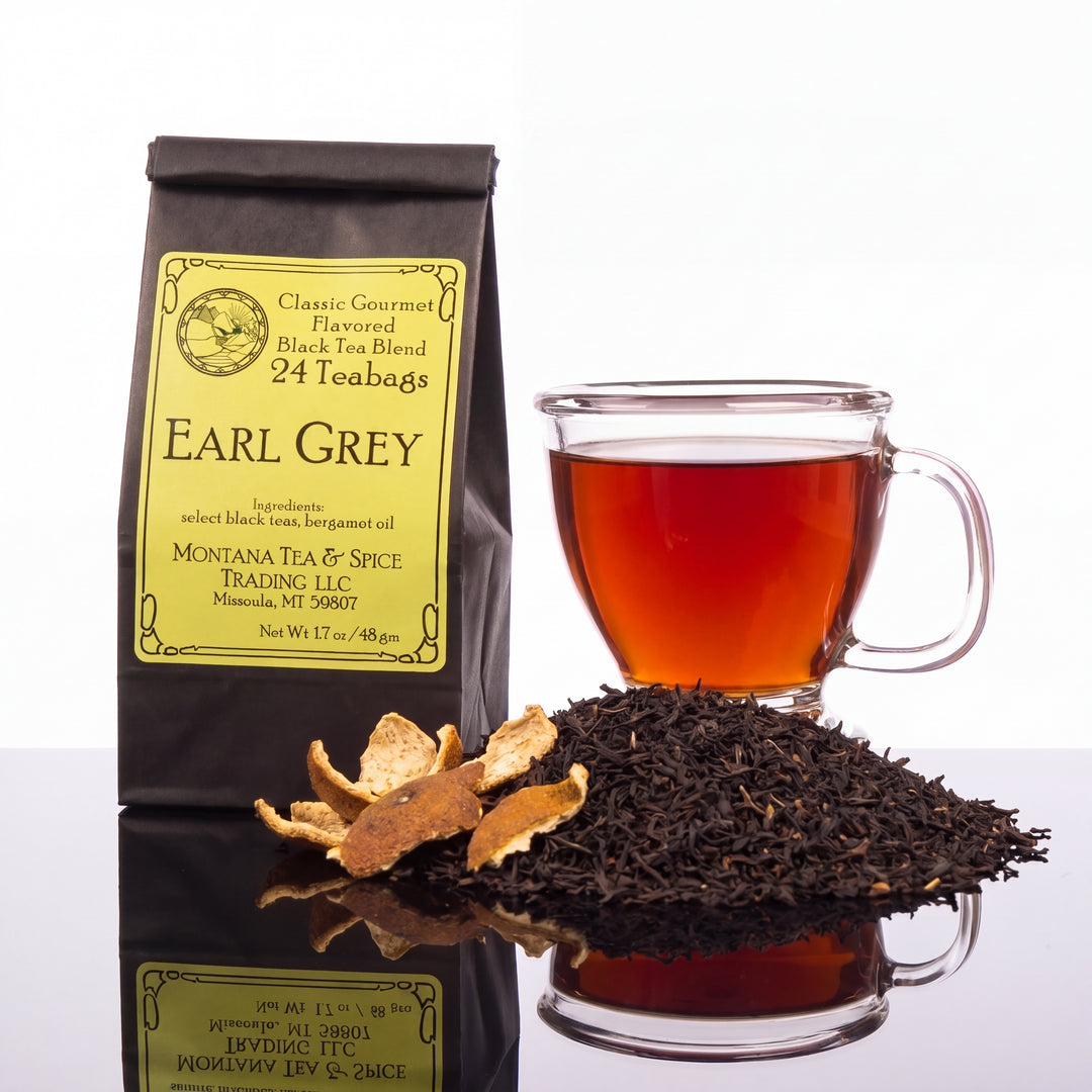 A bag of Montana Tea &amp; Spice Trading LLC Earl Grey tea stands next to a clear glass mug filled with amber tea, with loose black tea leaves and dried citrus slices in front on a white reflective surface.