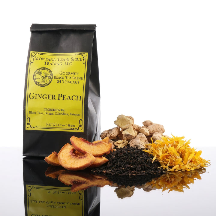 A black tea bag reading Ginger Peach from Montana Tea and Spice on a reflective surface with dried ginger, black tea, ginger root and dried peach slices